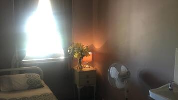 Una habitación de hotel con cama, mesita de noche con lámpara, ventilador y ventana con cortinas.
