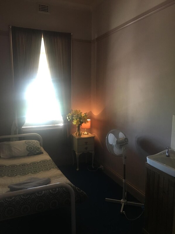 Una habitación de hotel con cama, mesita de noche con lámpara, ventilador y ventana con cortinas.