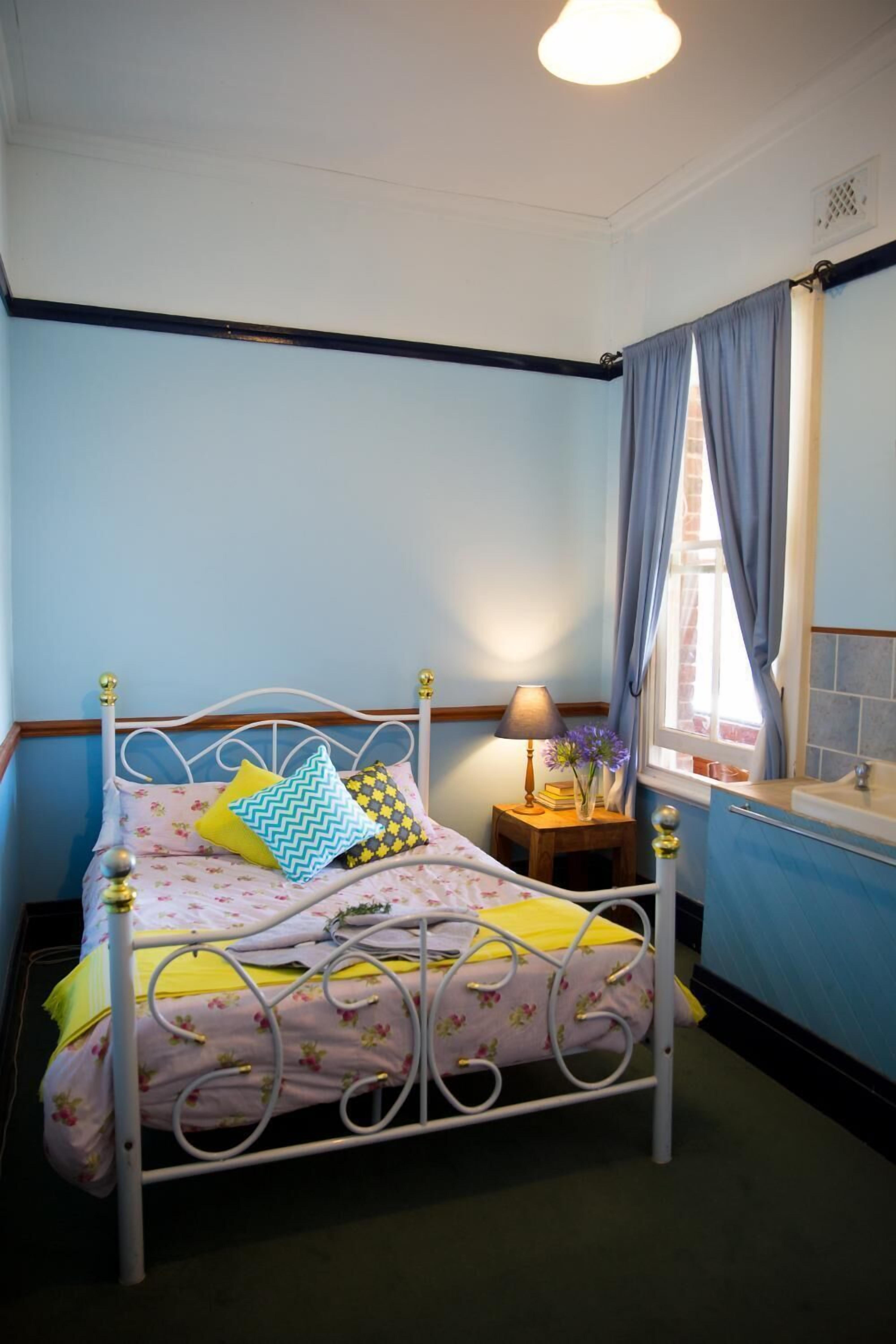 A bedroom with a bed, a bedside table with a lamp, a window with curtains, and a bathroom with a sink and mirror.