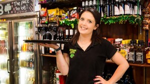 A person holding a tray with glasses, standing in a bar area with shelves of liquor bottles. - Boorowa Hotel (Boorowa)