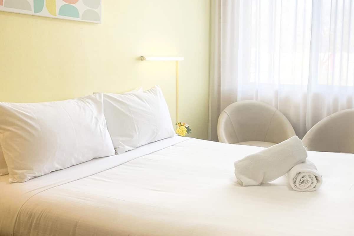 Ein Hotelzimmer mit einem ordentlich bezogenen Bett, zwei Stühlen, einer Lampe und einem Fenster mit transparenten Vorhängen.