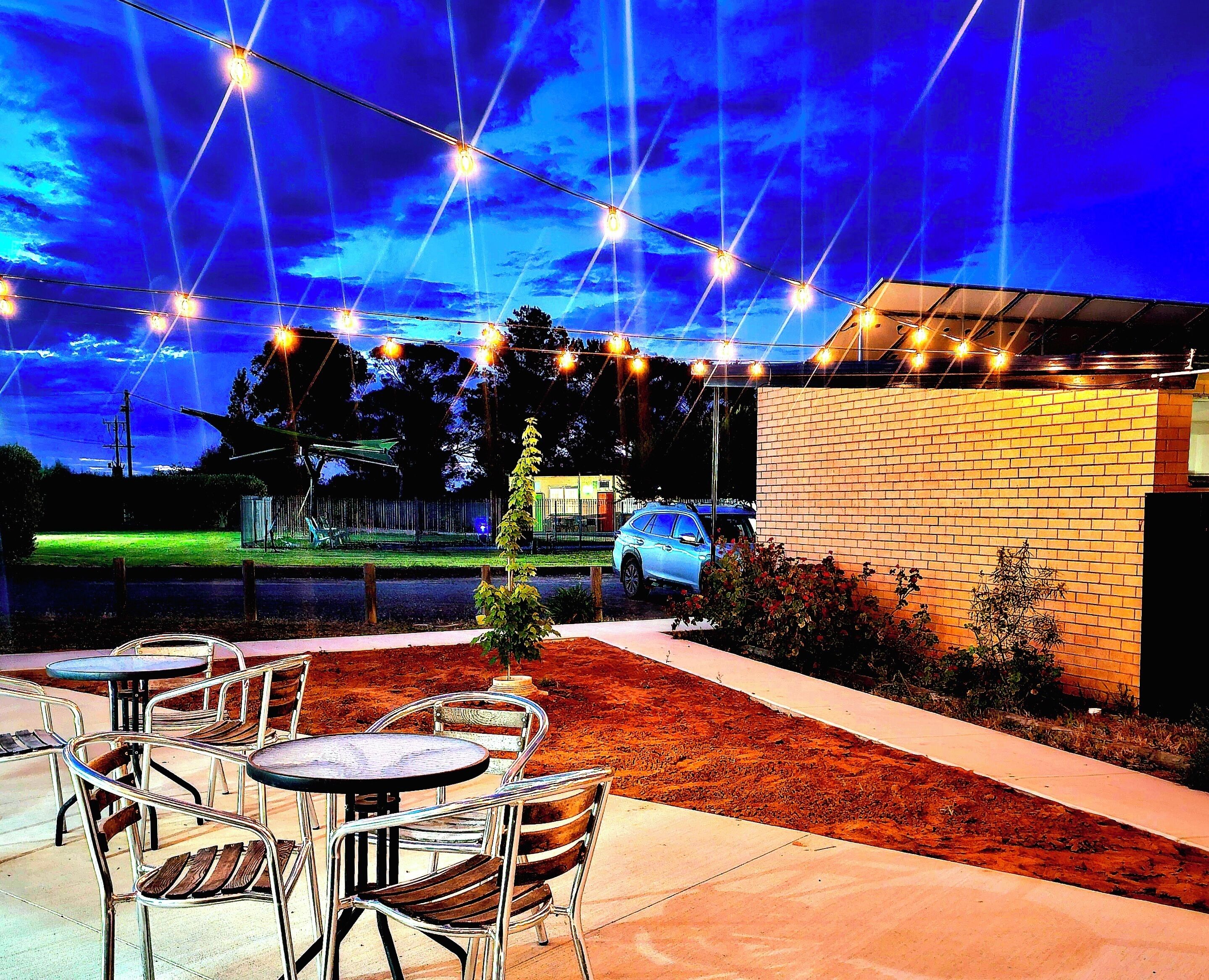 An outdoor patio area with tables and chairs, a brick wall, and a car parked in the background. - Sea Lake Motel (Sea Lake)