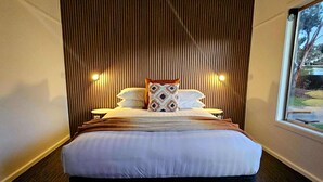 Una habitación de hotel moderna con una cama grande, cabecera de madera con listones, dos lámparas de noche y una ventana con vistas a árboles.