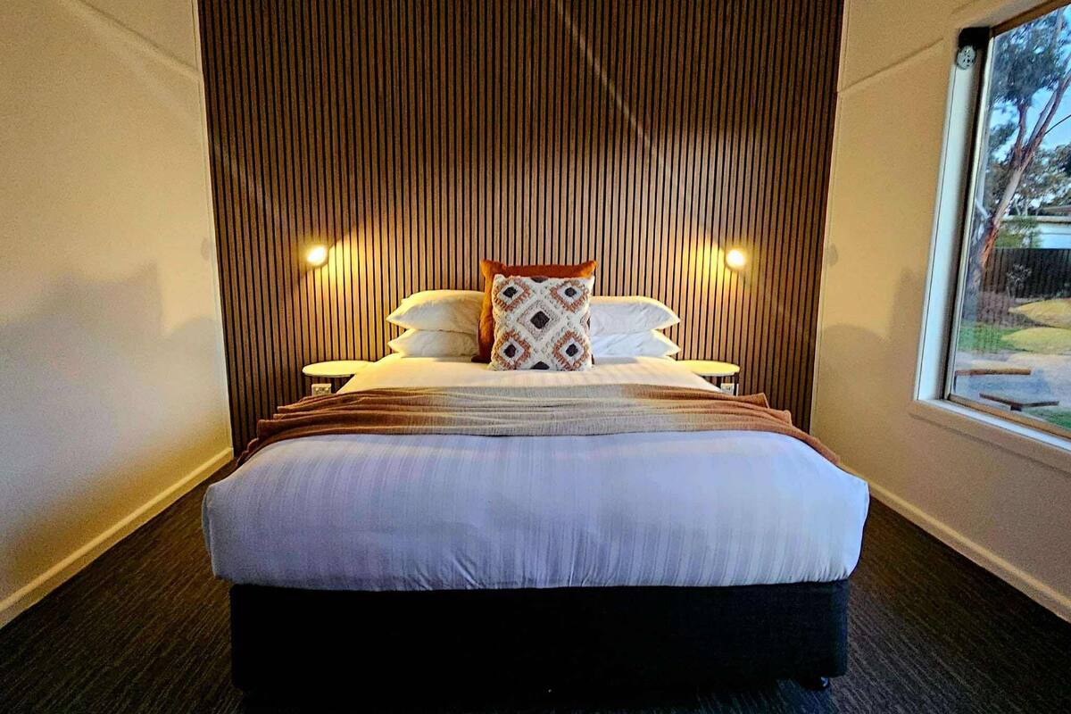 Ein modernes Hotelzimmer mit einem großen Bett, einem Holz-Schuppenverkleidungskopfstück, zwei Nachttischlampen und einem Fenster mit Blick auf Bäume.