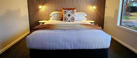 Una habitación de hotel moderna con una cama grande, cabecera de madera con listones, dos lámparas de noche y una ventana con vistas a árboles.