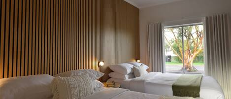 A hotel room with two beds, a wooden slat wall, a window with curtains, and a view of trees.