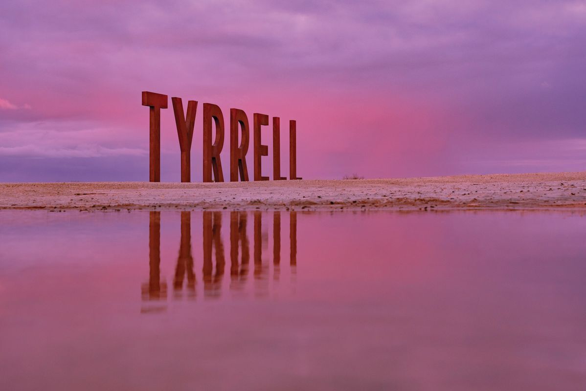 Large wooden letters spelling "TYRRELL" on a flat surface with a reflective water body in the foreground and a pinkish sky in the background.