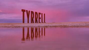 Large wooden letters spelling "TYRRELL" on a flat surface with a reflective water body in the foreground and a pinkish sky in the background.