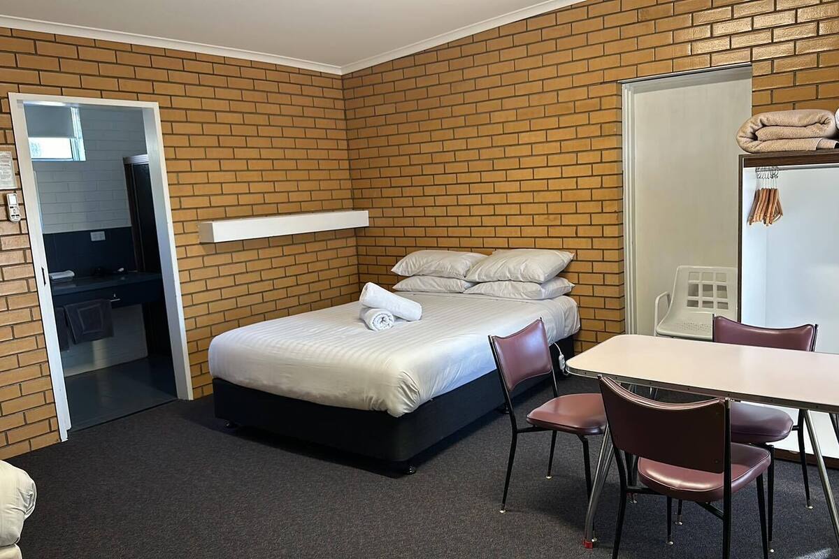 Ein kleines Hotelzimmer mit Backsteinwand, einem Bett mit weißen Laken, einem kleinen Tisch mit Stühlen und einem Kleiderschrank mit Handtüchern.