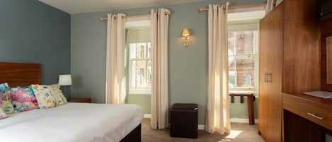 A bedroom with a bed, a nightstand, a lamp, a wooden wardrobe, and a window with curtains.