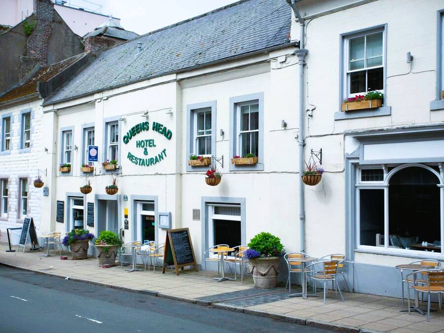 A white building with a sign reading "Queens Head Hotel & Restaurant". There are outdoor tables and chairs, and hanging flower baskets.