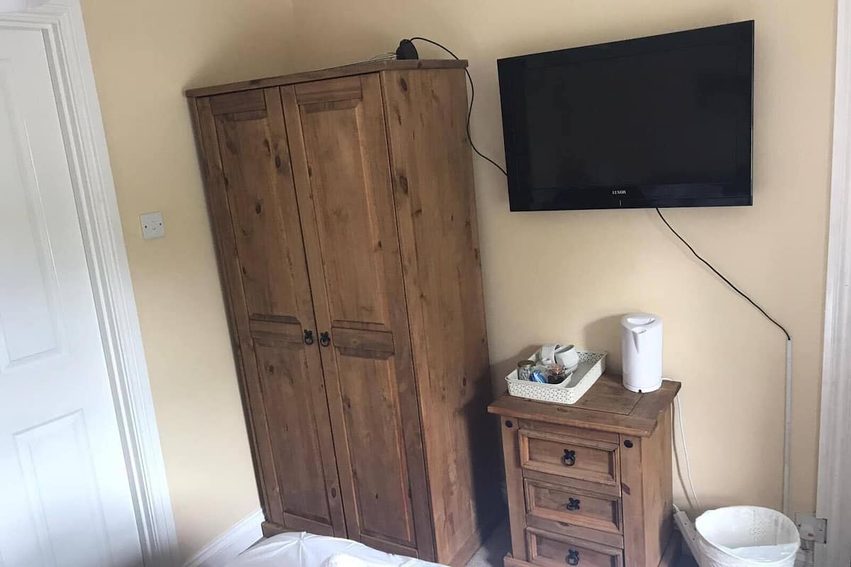 A bedroom with a wooden wardrobe, a small wooden dresser, a television mounted on the wall, and a white trash bin.