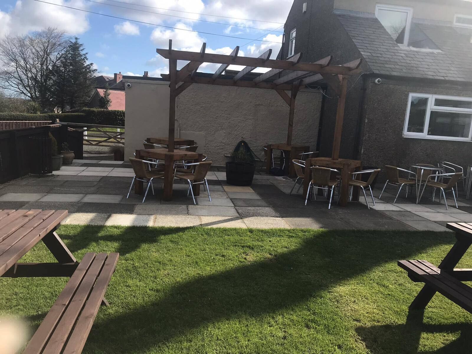 An outdoor patio area with wooden tables and chairs, a pergola, and a grassy lawn.