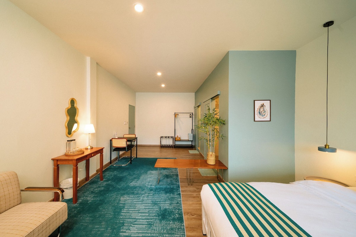 Habitación de hotel con cama, escritorio, silla, mesita, lámpara, planta y espejo.