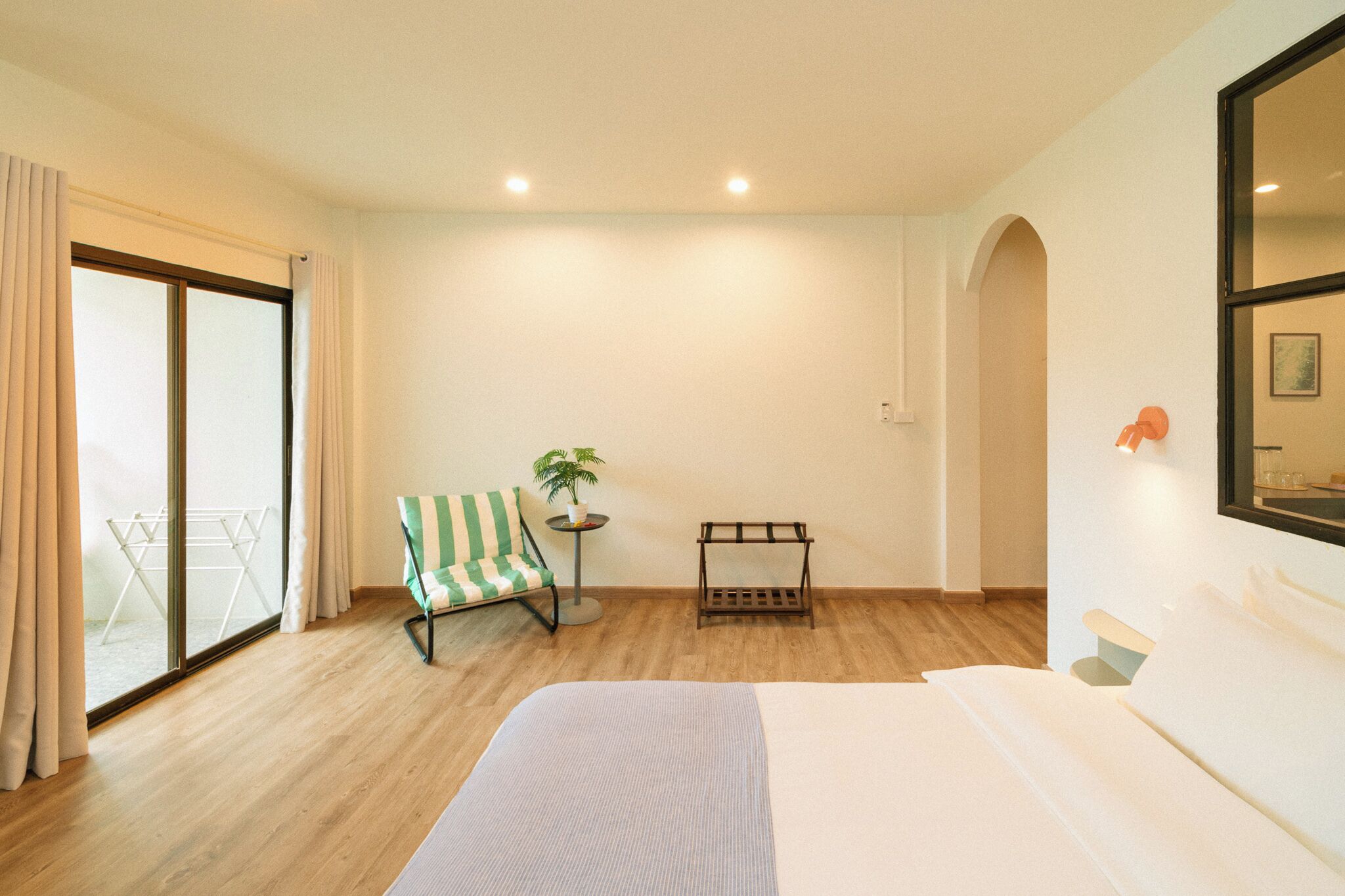 A hotel room with a bed, a chair, a small table with a plant, a drying rack, and a sliding glass door leading to an outdoor area.