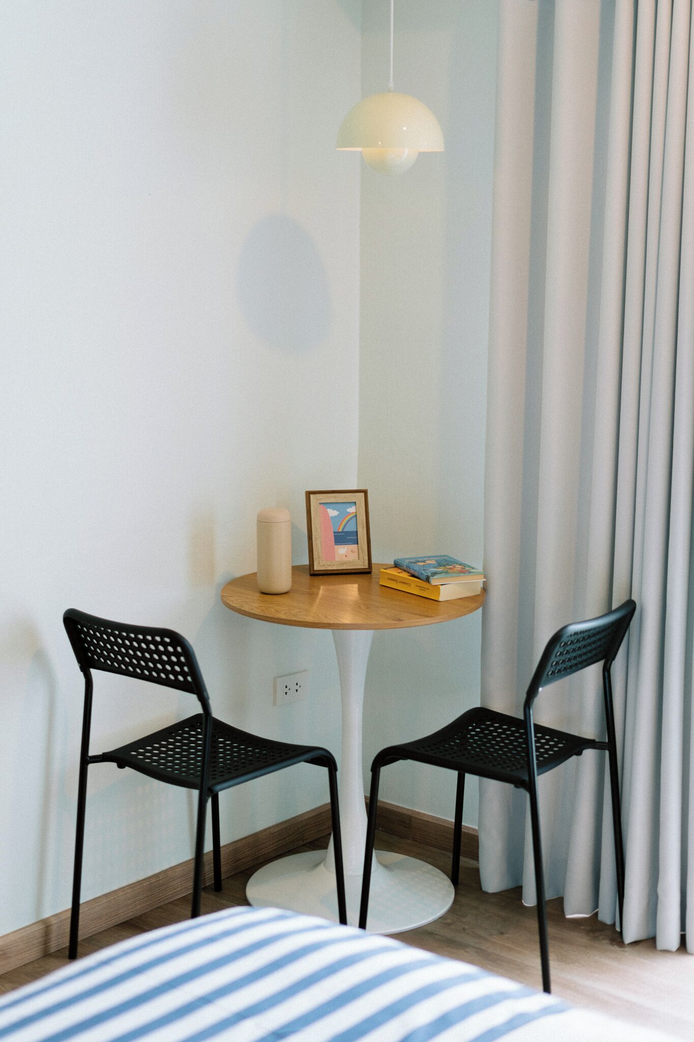 A small, neatly arranged room with a round wooden table, two black chairs, a single hanging light fixture, a framed picture, and a few books.