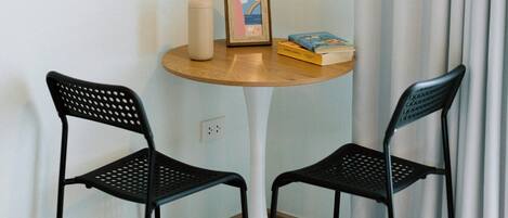 A small, neatly arranged room with a round wooden table, two black chairs, a single hanging light fixture, a framed picture, and a few books.
