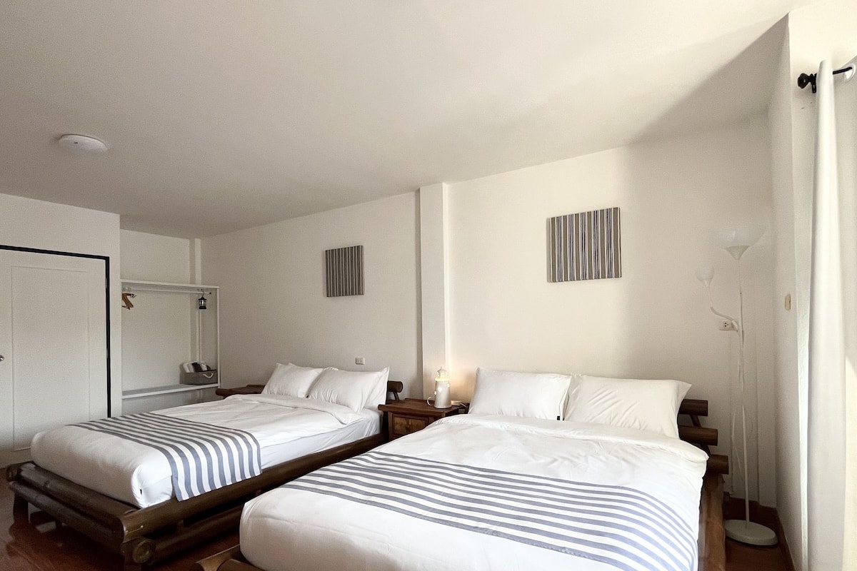 Habitación de hotel con dos camas, piso de madera, puerta blanca y una mesita de noche con lámpara.
