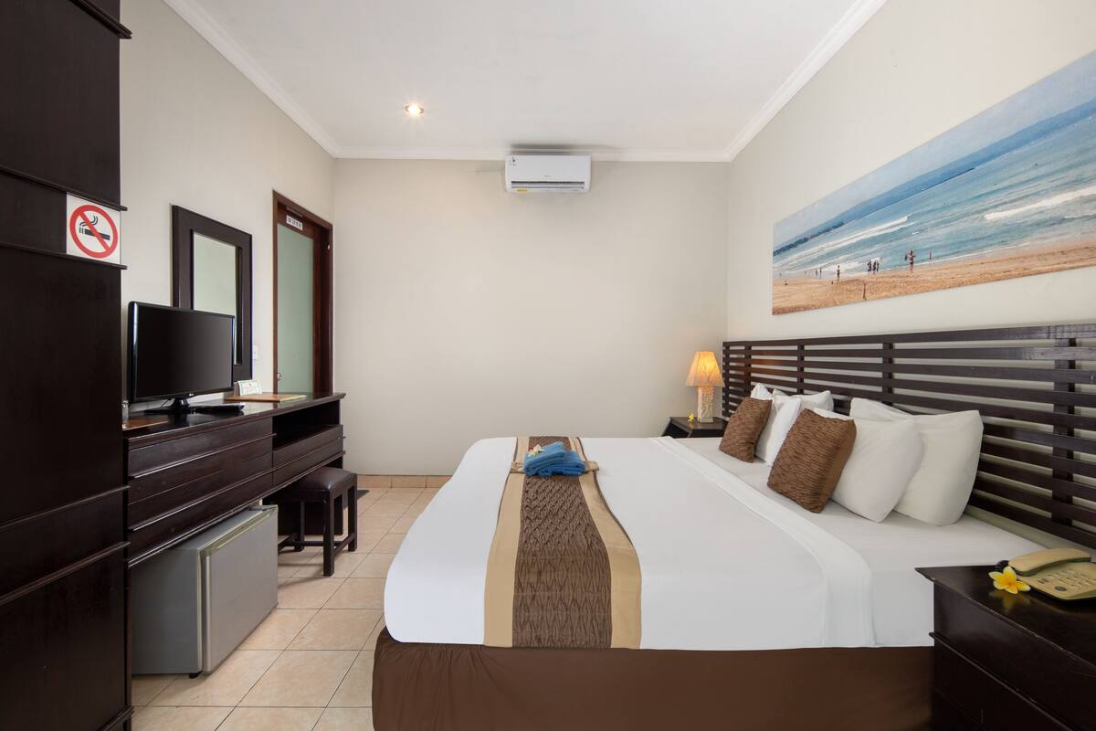 A hotel room with a bed, a desk with a computer, a TV, and a painting of a beach.