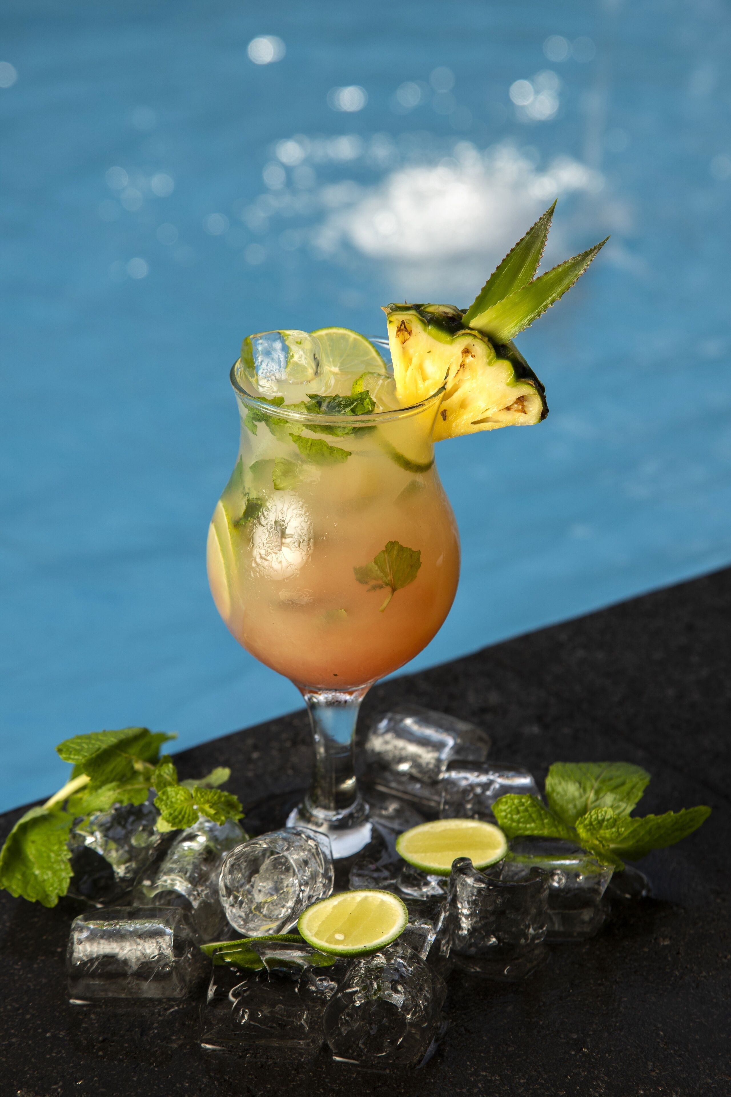 A cocktail with a pineapple garnish, mint leaves, and lime slices, served in a glass with ice cubes, placed on a dark surface with a pool in the background.