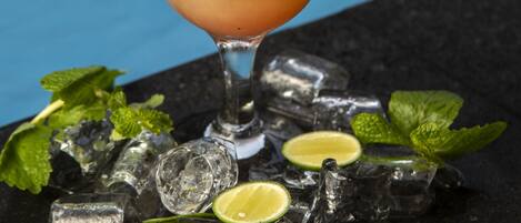 A cocktail with a pineapple garnish, mint leaves, and lime slices, served in a glass with ice cubes, placed on a dark surface with a pool in the background.