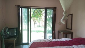 A bedroom with a bed, a standing fan, a table, and a window with curtains. - Pai Park Cottage (Pai)