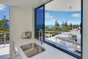 A modern kitchen with a large window overlooking a scenic view of the ocean and trees. - Breeze Mooloolaba (Mooloolaba)