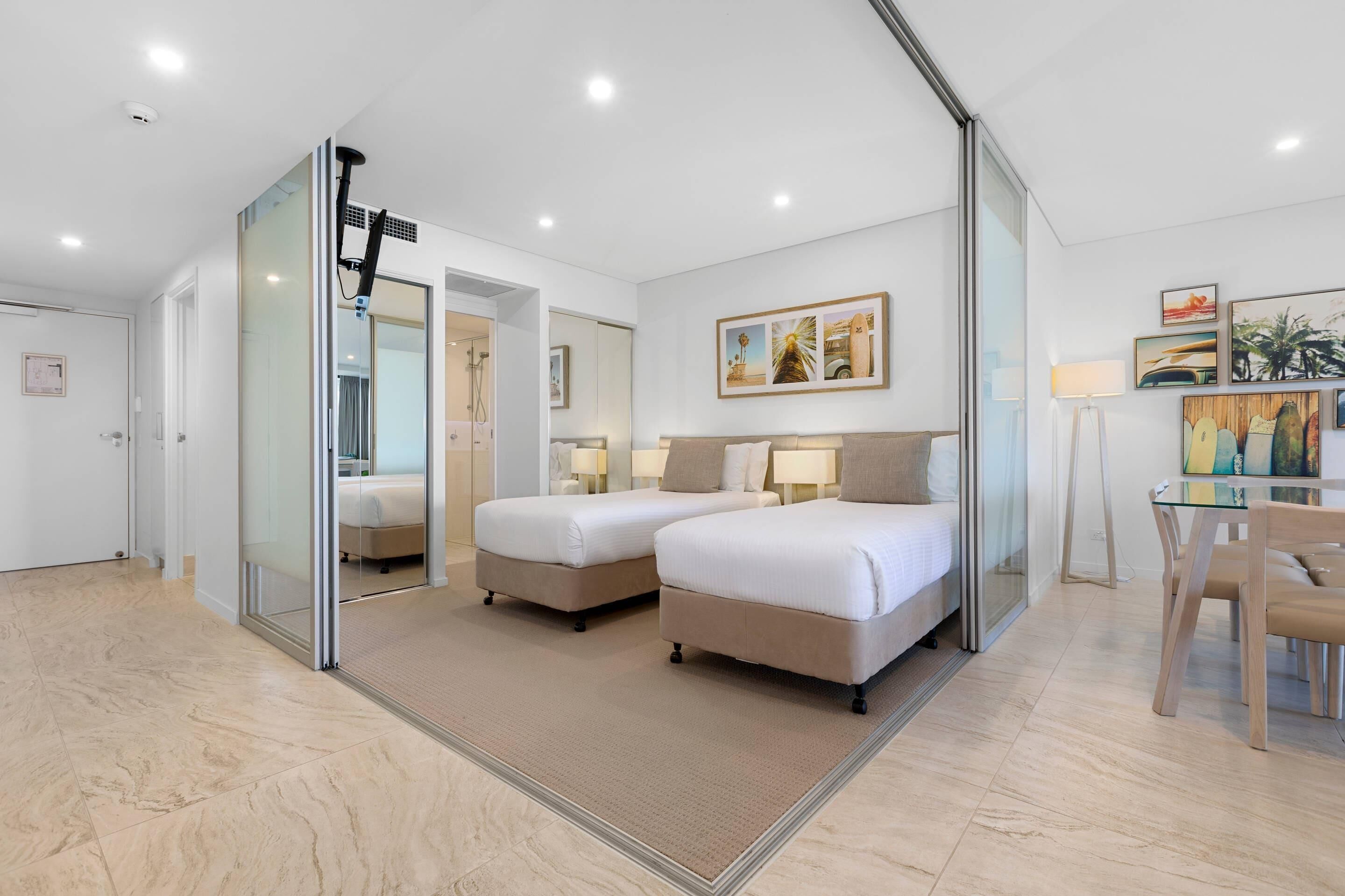 Una habitación de hotel moderna con dos camas, cabina de ducha de vidrio, zona de comedor con mesa y sillas, y obras de arte en las paredes.