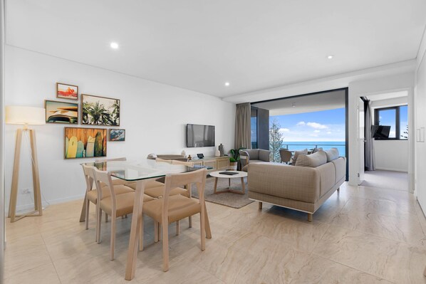 A modern living room with a glass dining table, beige sofa, and a large window offering a view of the sea. - Breeze Mooloolaba (Mooloolaba)