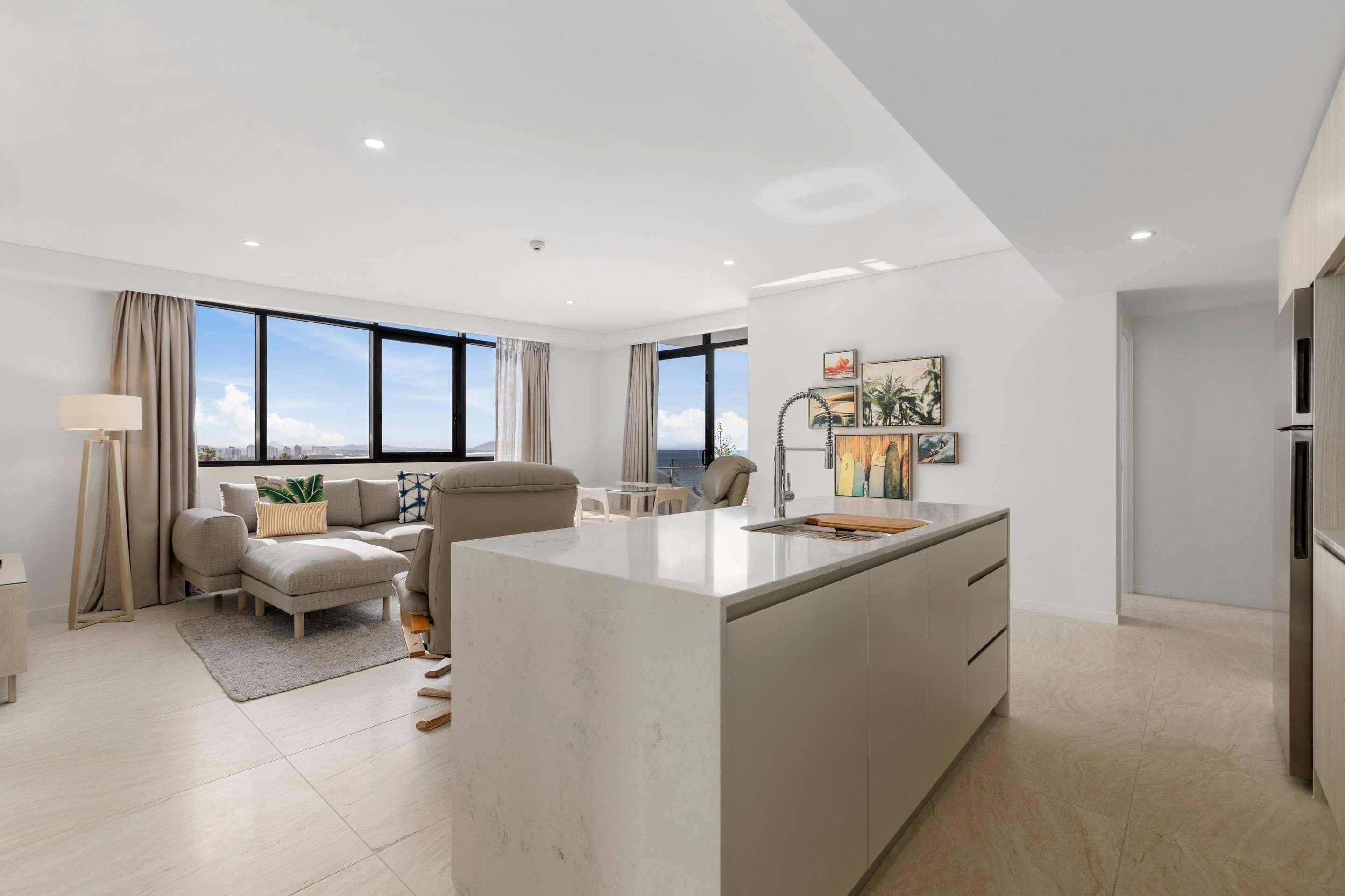 A modern living space with a large island kitchen, a sofa set, a dining table, and a view of the city.