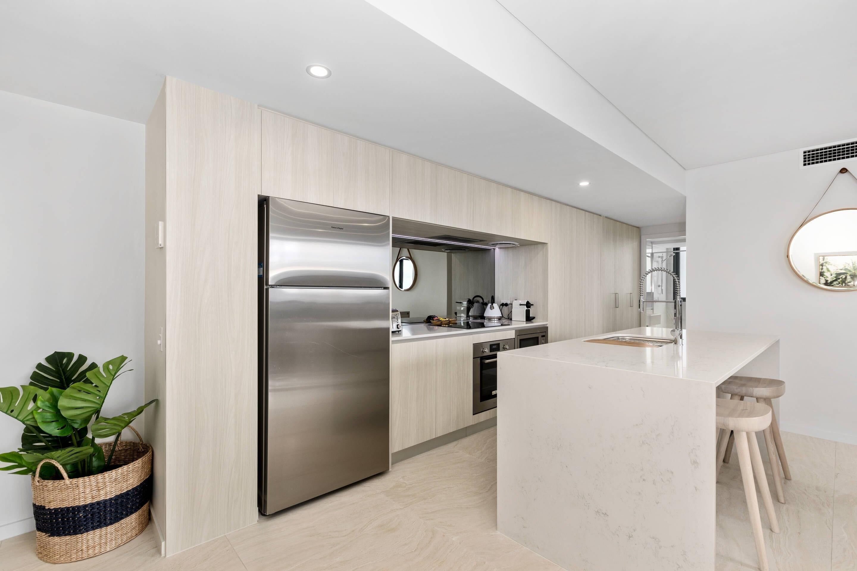 A modern kitchen with a stainless steel refrigerator, wooden cabinets, a marble countertop, and a plant.