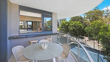 A balcony with a table and chairs, a window with a view of buildings and trees, and a railing.