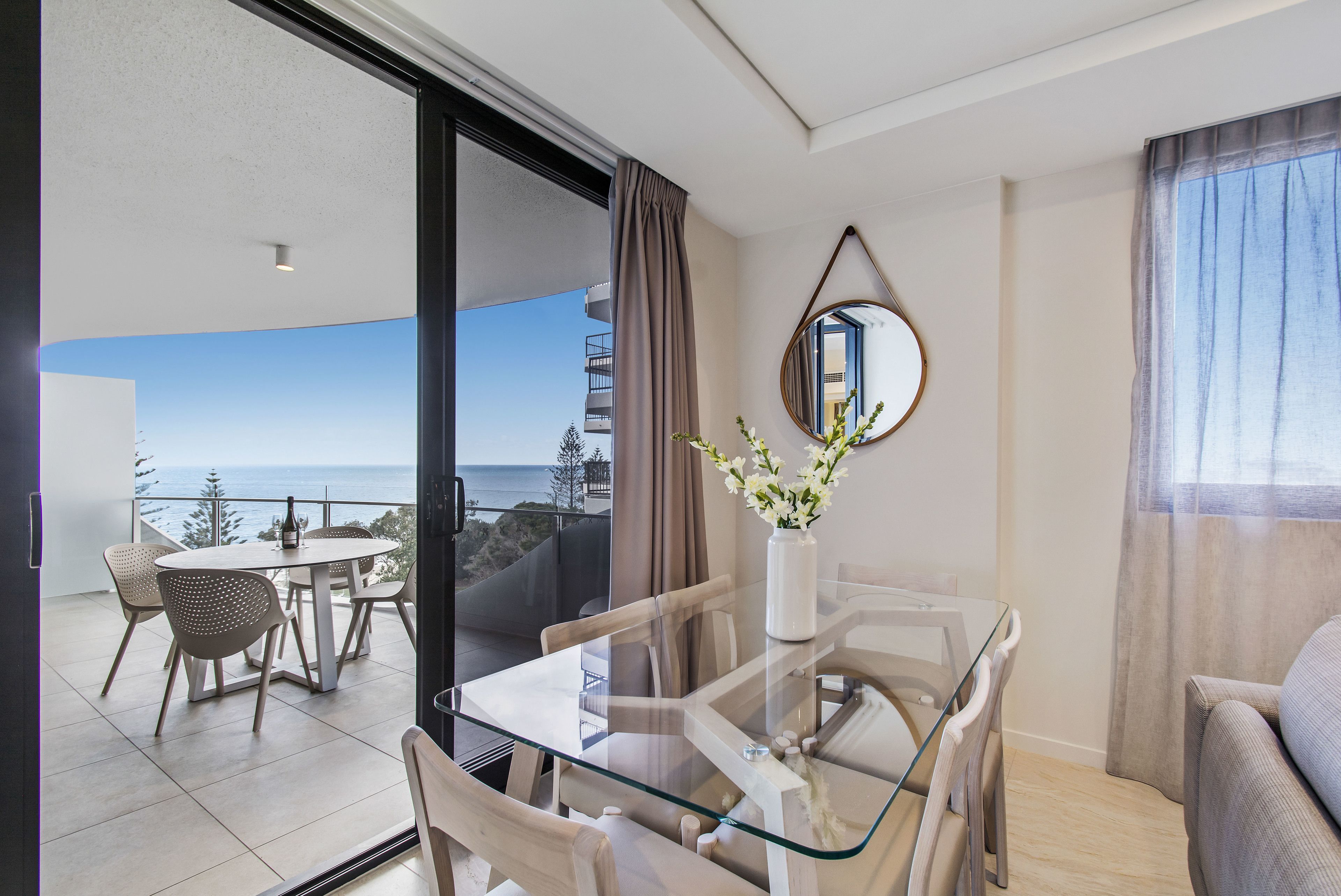A modern living room with a glass dining table, a sofa, and a view of the ocean.