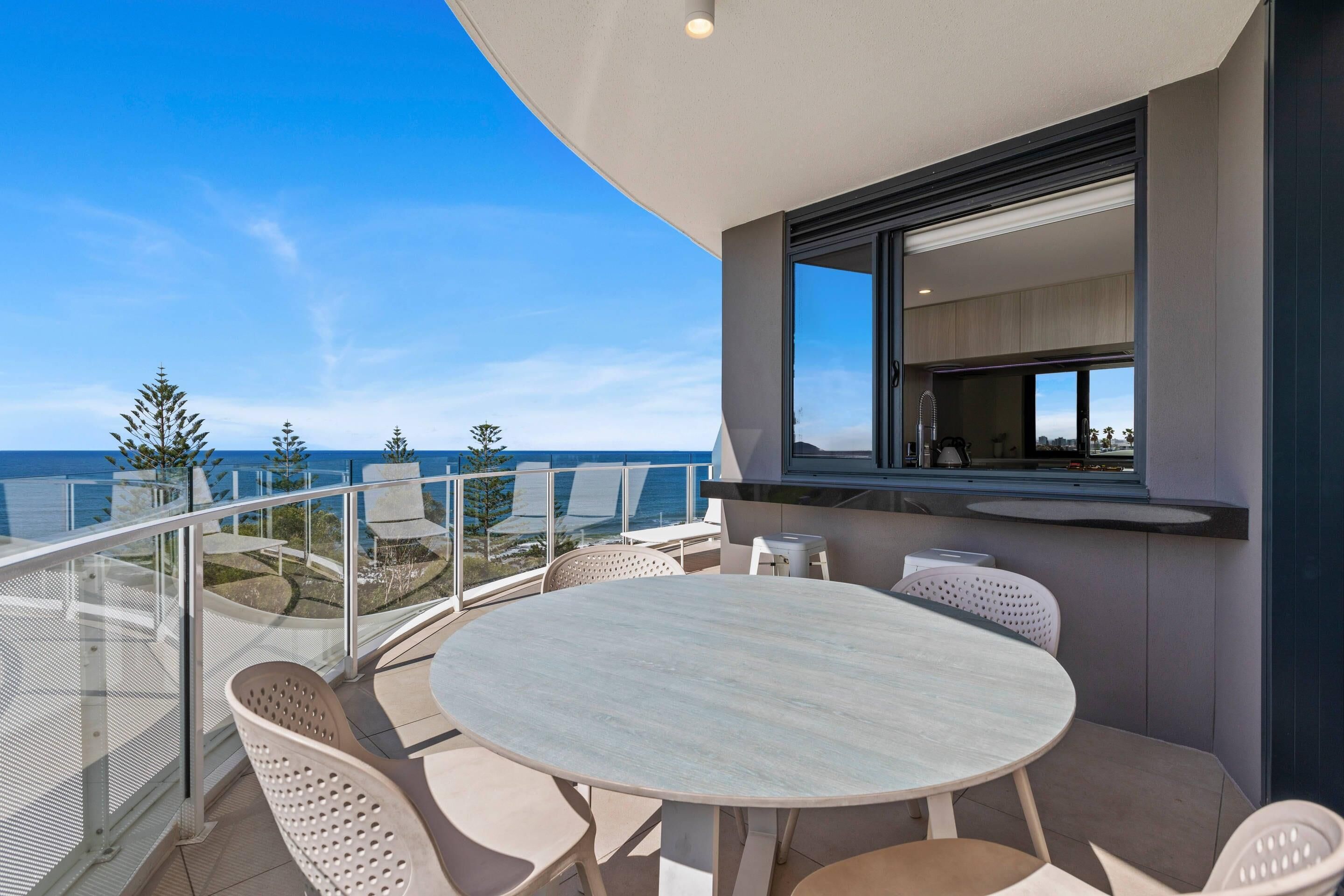 A balcony with a round table and chairs, a glass railing, and a view of the sea.