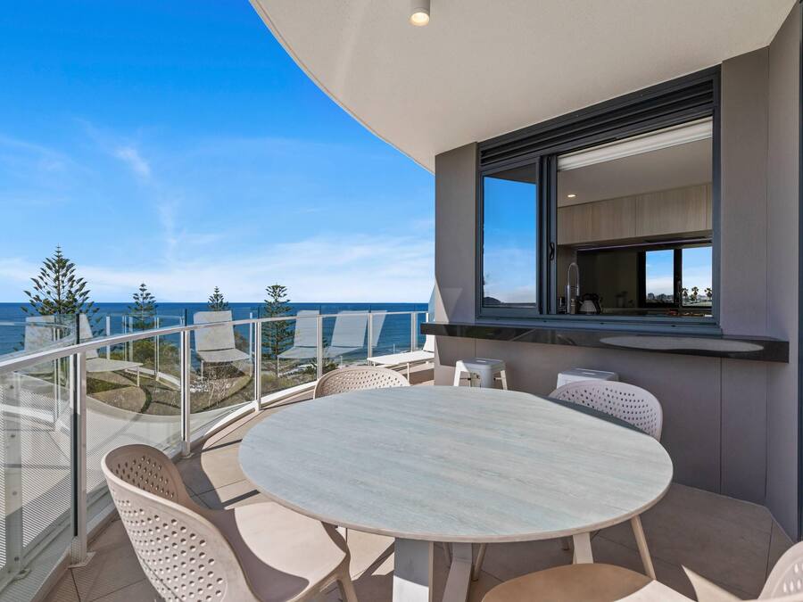 A balcony with a round table and chairs, a glass railing, and a view of the sea.