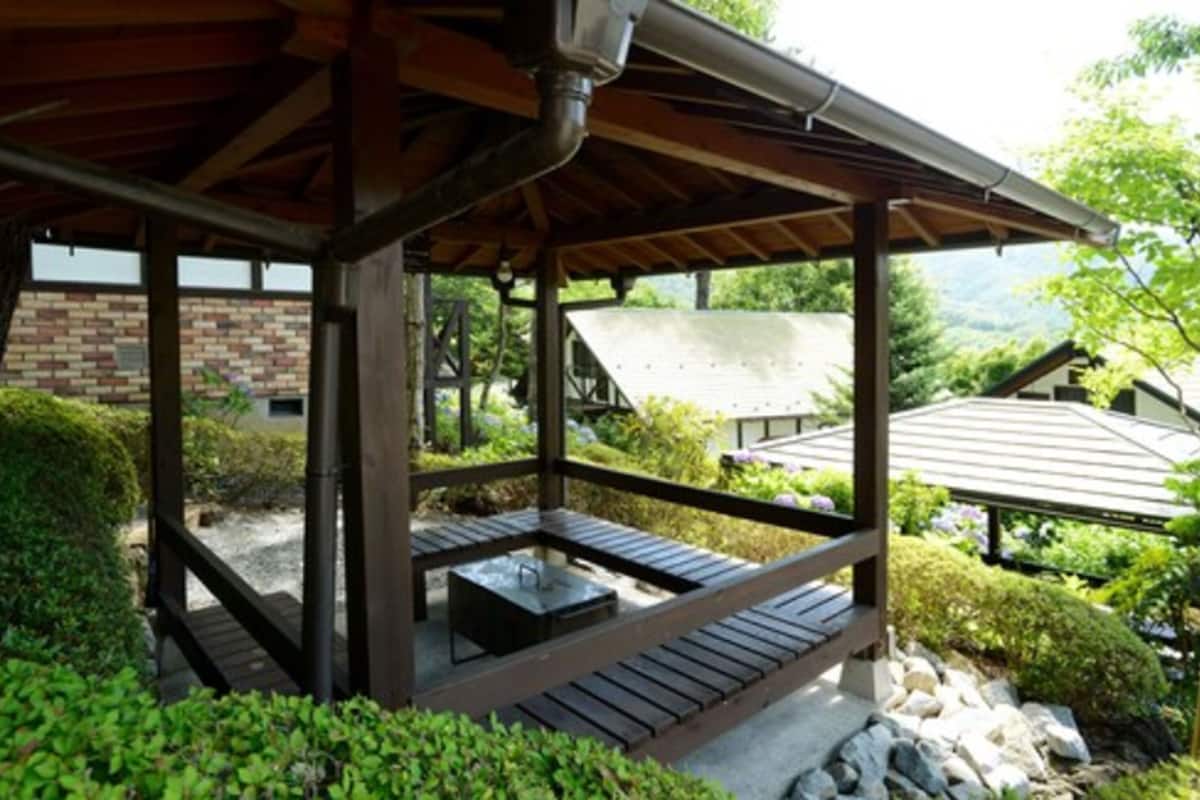 A covered outdoor seating area with a table and benches, surrounded by greenery and a view of a building in the background.