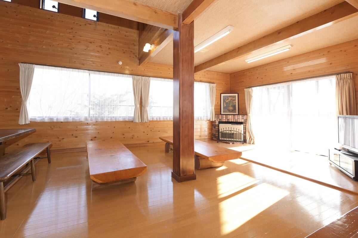 A spacious room with wooden floors and walls, large windows with curtains, a wooden bench, and a television.