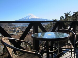 A balcony with wicker chairs and a glass table overlooking a town and a snow-capped mountain. - Kawaguchiko country cottage Ban (Fujikawaguchiko)
