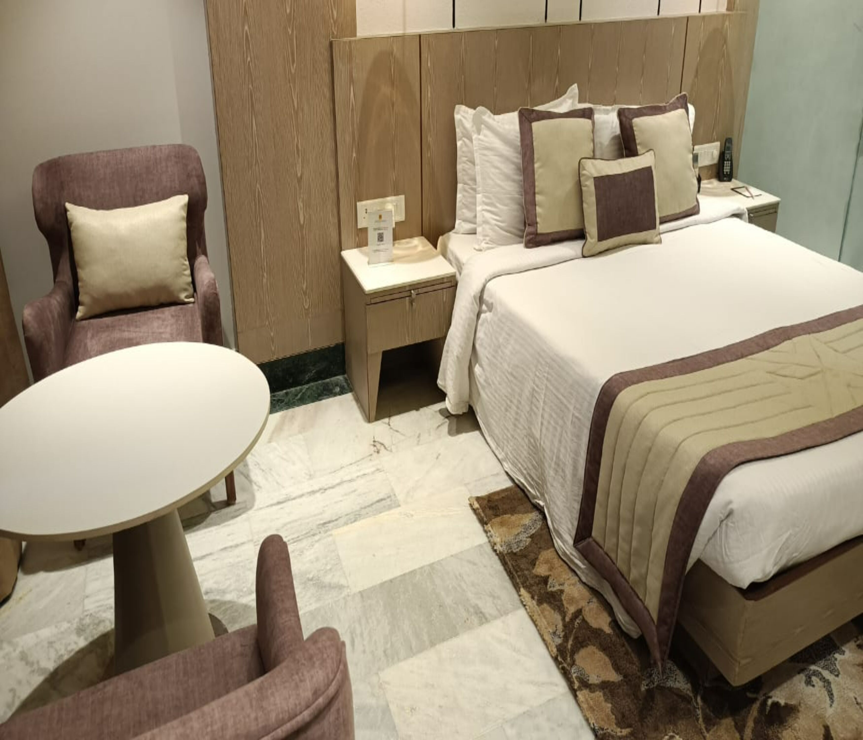 Quarto de hotel com cama, uma cadeira, uma mesinha e um criado-mudo.