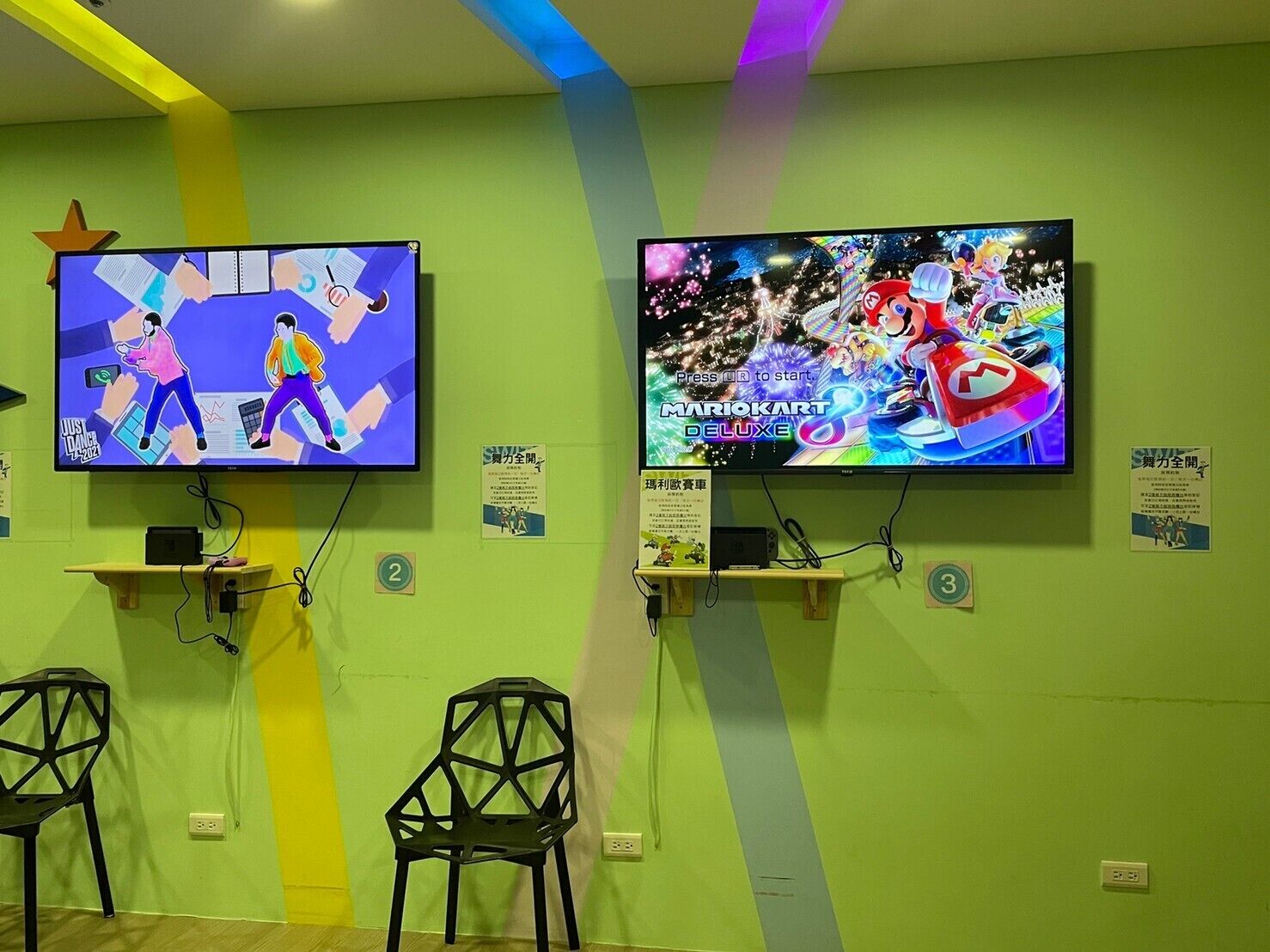Two televisions mounted on a green wall, each displaying different video games. Below the TVs, there are two black chairs and a small shelf with a book.