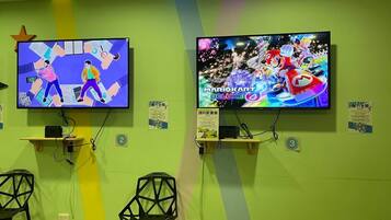 Two televisions mounted on a green wall, each displaying different video games. Below the TVs, there are two black chairs and a small shelf with a book.
