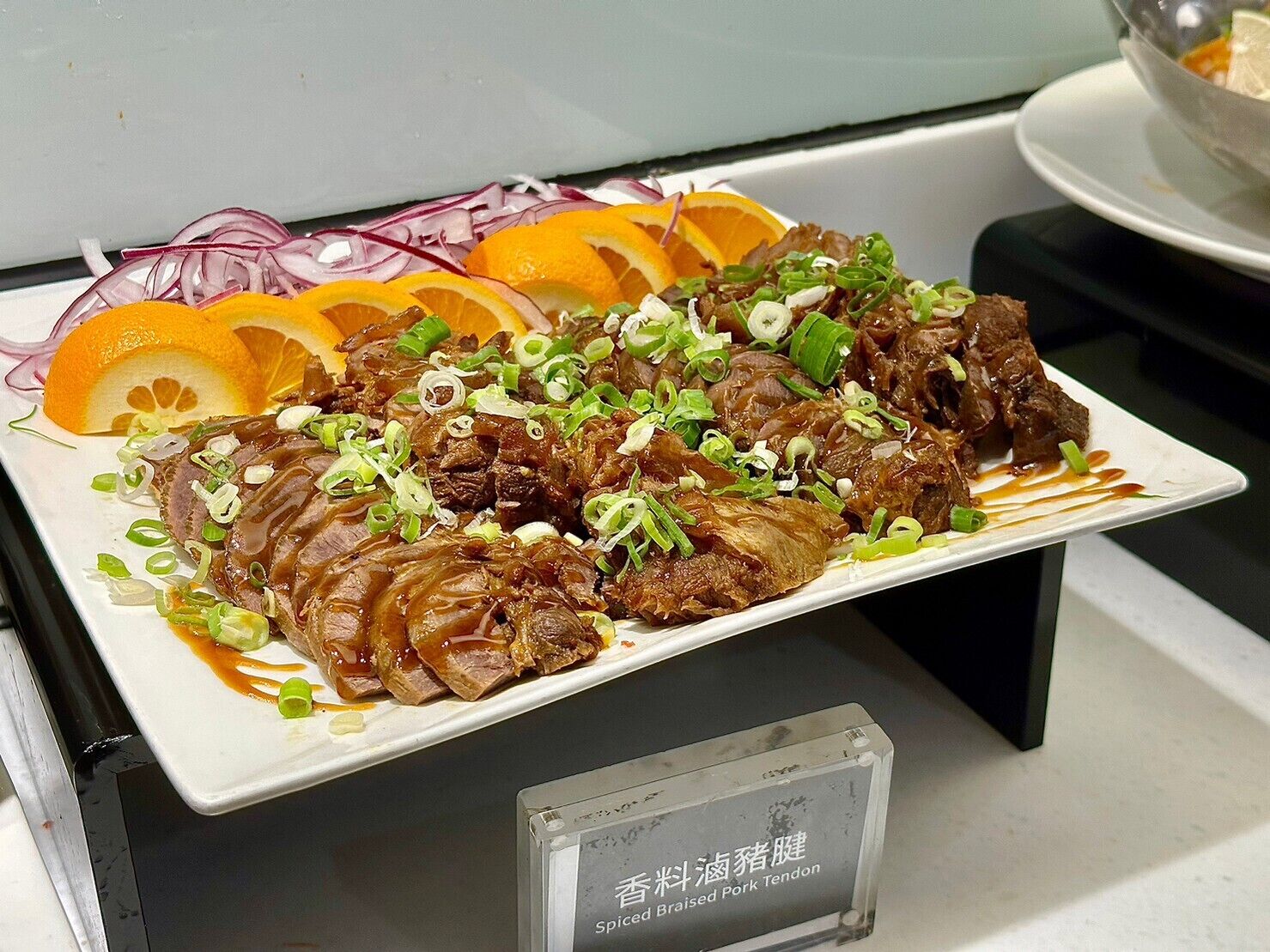 A plate of braised pork tendon garnished with green onions and orange slices, with a label indicating it is spicy.