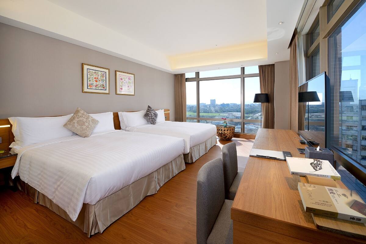 A hotel room with two beds, a desk, and a large window offering a city view.