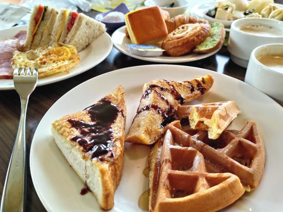 A breakfast plate with waffles and a slice of cake, accompanied by a sandwich and a croissant.