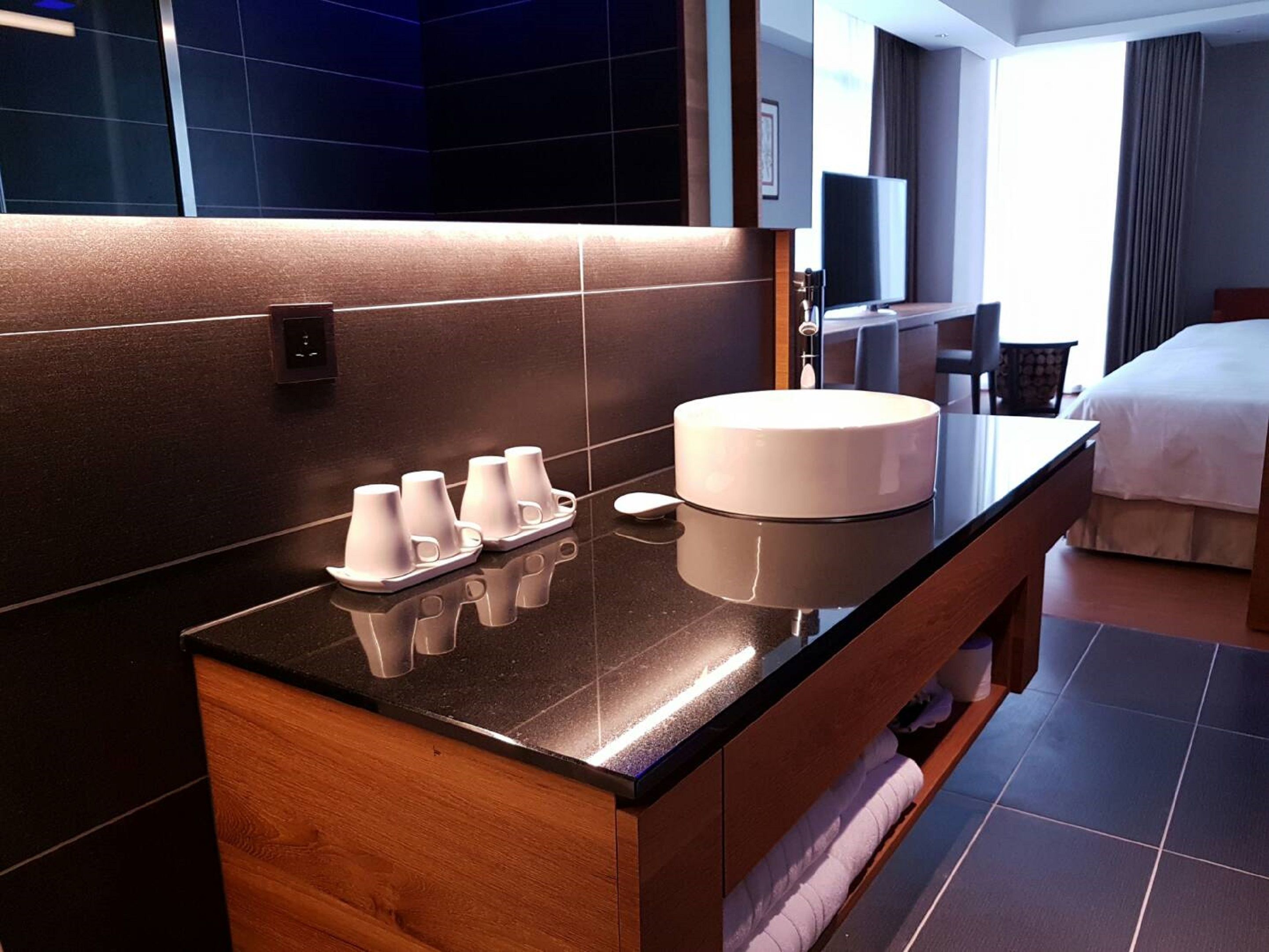 A modern hotel bathroom with a large mirror, a round sink, and a tray of white cups.