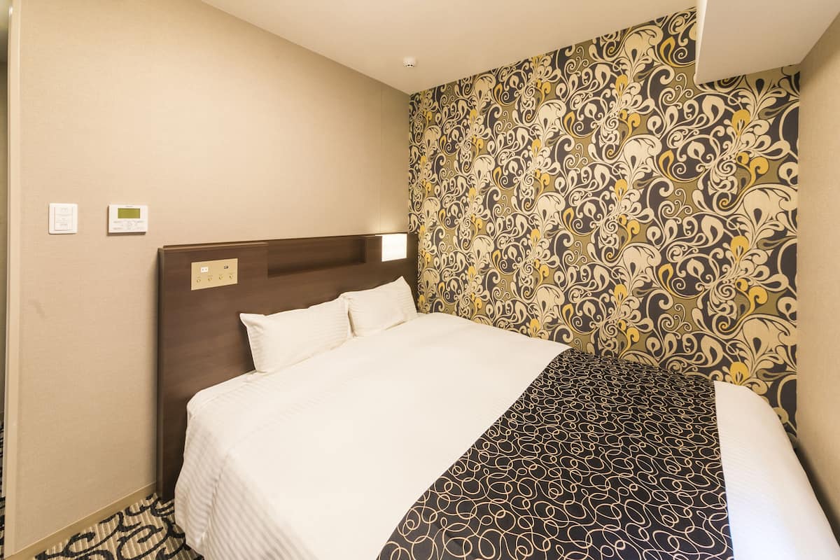 Quarto de hotel com uma cama grande, cabeceira, papel de parede estampado e painel de controle na parede.