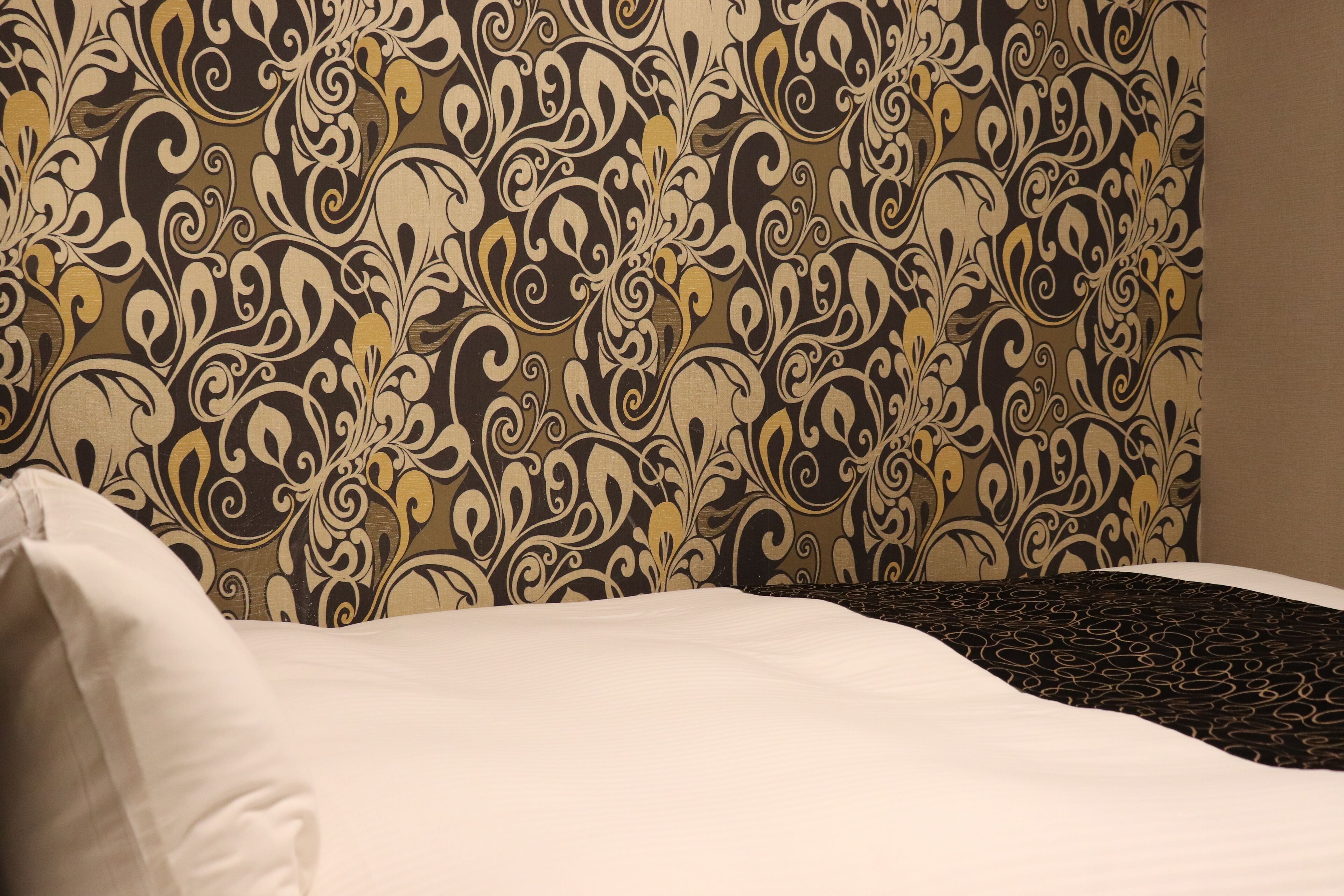 A hotel room with a patterned wallpaper, a neatly made bed, and a dark patterned throw on the bed.