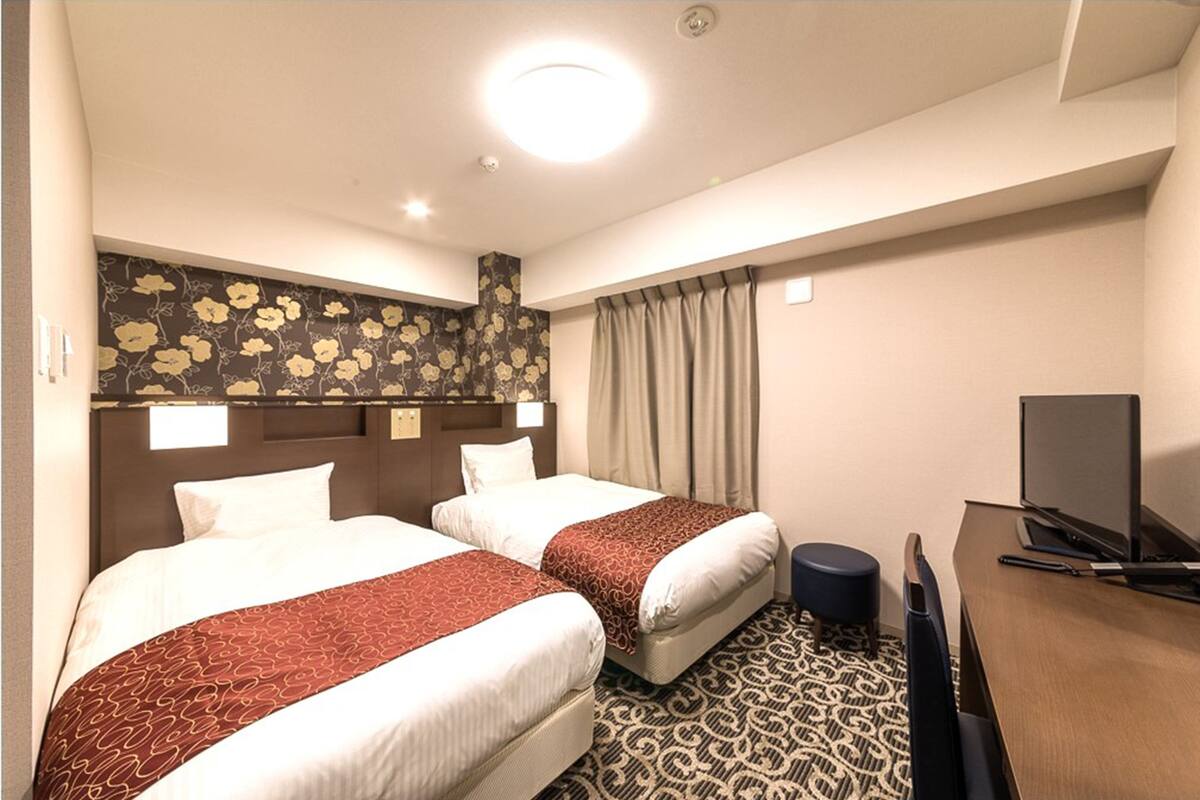 Quarto de hotel com duas camas, uma escrivaninha, televisão e carpete estampado.