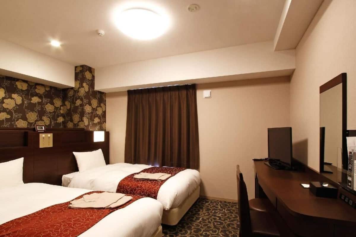 Quarto de hotel com duas camas, uma escrivaninha, uma cadeira, um espelho e um tapete estampado.