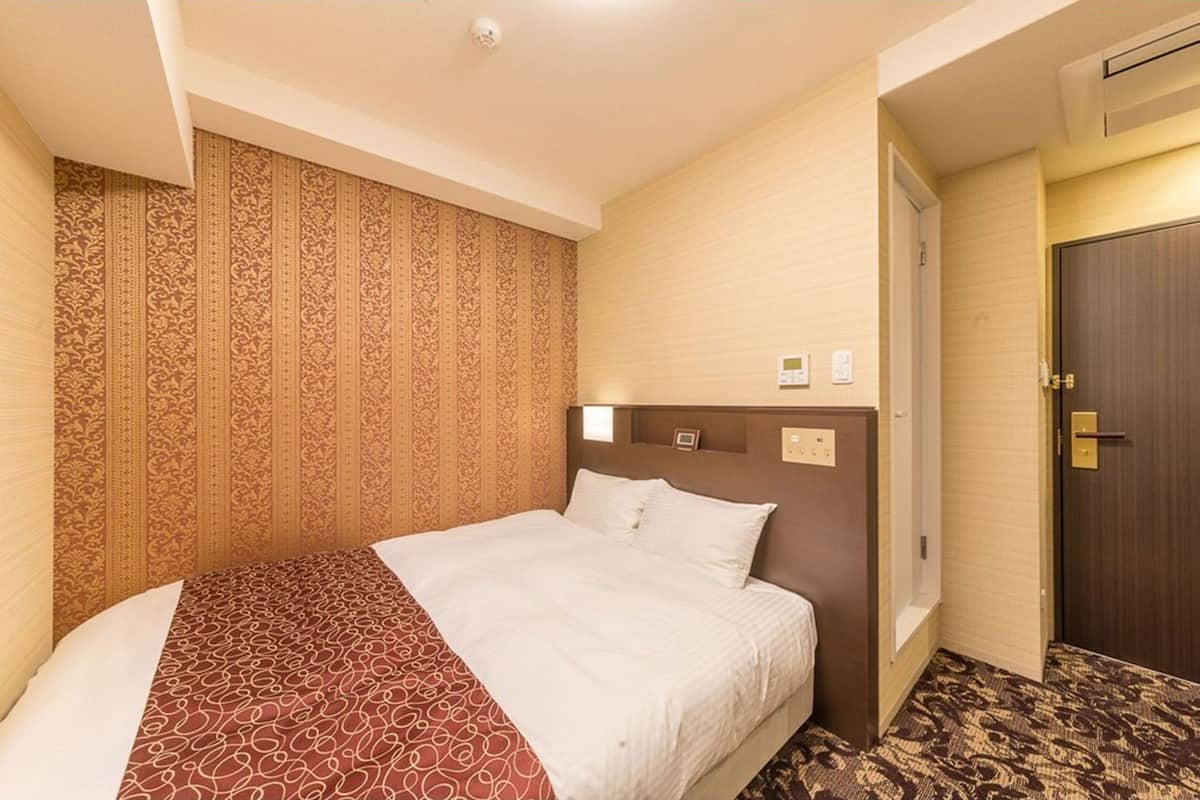 Quarto de hotel com uma cama grande, papel de parede estampado, uma porta de madeira e um interruptor de luz na parede.